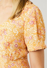 Blusa amarilla con flores que presenta mangas cortas abullonadas, patrones florales en blanco y rosa, y una cintura fruncida con un dobladillo con volantes en las mangas.