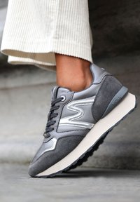 Sneaker grigio caratterizzato da una combinazione di materiali lisci e testurizzati, forma arrotondata, lacci e un design con accenti argentati sui lati.