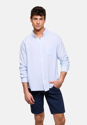 Hombre de pie con camisa de manga larga azul claro abotonada y pantalones cortos azul marino, con una mano en el bolsillo contra un fondo liso.