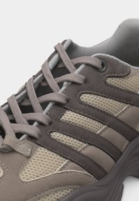 Chaussure de sport beige et marron foncé avec tissu en mesh, empiècements en daim et lacets épais assortis noués sur la languette.