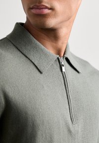 Pullover verde chiaro, texturizzato, con colletto con zip, caratterizzato da un collo aderente e dettagli cuciti lungo i bordi del colletto.