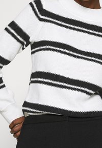 Gestreifter Strickpullover in Weiß und Schwarz, mit breiten horizontalen Streifen, gerippten Bündchen und rundem Halsausschnitt. Texturierte Stoffdetails sichtbar.