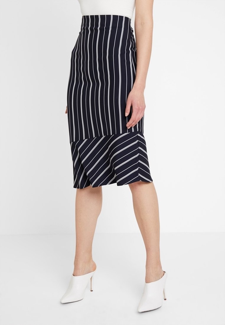 Jupe crayon bleu marine à rayures, avec un ourlet asymétrique, présentant des rayures blanches verticales et diagonales, et une silhouette ajustée.