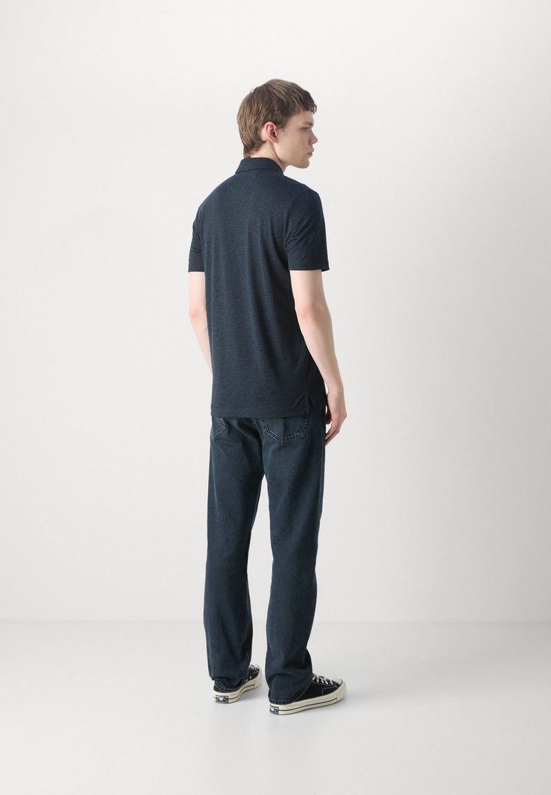 Dunkelblaues Poloshirt aus Strickstoff, mit kurzen Ärmeln und klassischem Kragen, kombiniert mit dunklen Jeans und schwarzen Sneakers.