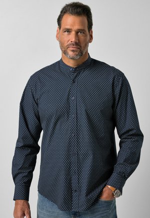 Mann mit kurzen dunklen Haaren und Bart, der ein dunkelblau gemustertes Hemd mit Knöpfen und hellblaue Jeans mit Armbanduhr trägt, steht vor einem einfarbigen Hintergrund.