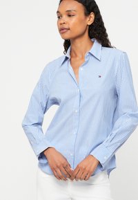 Blue and white striped button-up shirt with a collar, featuring long sleeves, a curved hem, and a small logo accent on the chest.