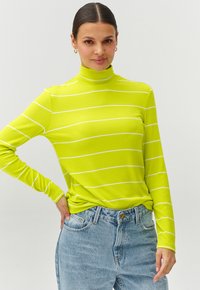Svetlo zelena rebrasta turtleneck majica z belimi horizontalnimi črtami, dolgimi rokavi in sproščenim krojem, v kombinaciji s svetlo modrimi visokim pasom kavbojkami.