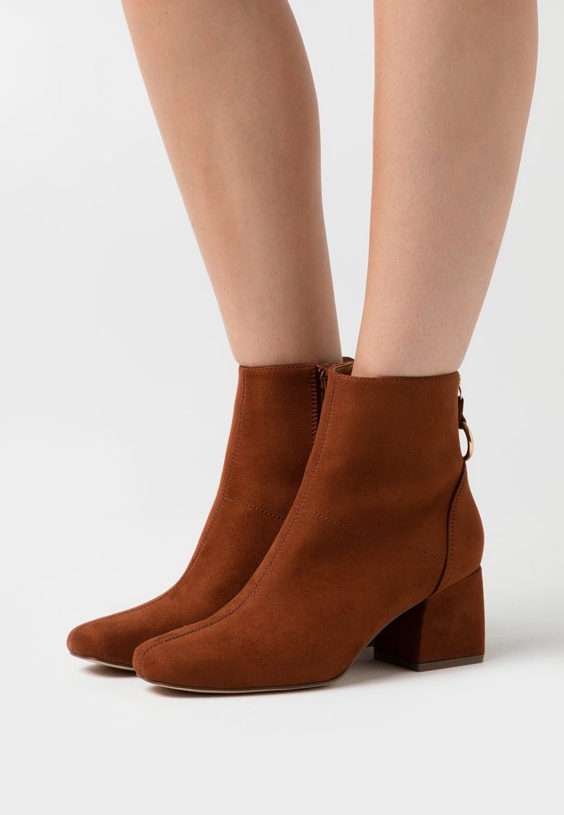 Bottines en daim marron à talons blocs portées sur des jambes nues sur fond blanc.
