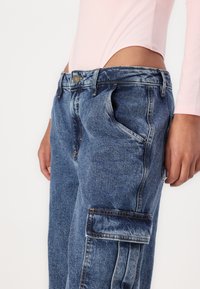 Visokotaknjen modri denim cargo hlače z sproščenim krojem, ki imajo več žepov in zbledelo teksturo. Podroben šiv ob šivih.