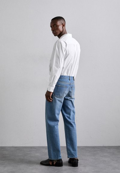 Jeans de mezclilla azul claro de pierna recta, camisa blanca de botones, zapatos negros con hebilla. Textura suave, corte casual, bolsillos traseros visibles.