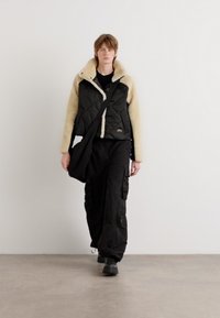 Κουβερτίνα μαύρη ζακέτα με κρέμ ψεύτικο λαιμό, συνδυασμένη με μαύρα cargo παντελόνια και μαύρη τσάντα χιαστί. Απλός, μίνιμαλ σχεδιασμός.