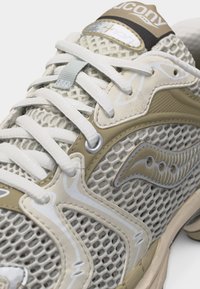 Αθλητικό παπούτσι Saucony σε μπεζ και λευκό χρώμα με ύφασμα από δίχτυ, λευκά κορδόνια και εμφανές λογότυπο στο πλάι και στη γλώσσα.