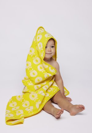 Malček sedi na tleh, ovit v rumeno brisačo z belimi krožnimi vzorci, z mirnim izrazom rahlo obrne pogled na stran.