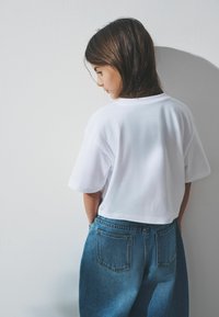 Weißes, kurzes T-Shirt mit kurzen Ärmeln, kombiniert mit hochgeschnittenen blauen Jeans aus Denim, die hintere Taschen und einen lockeren Schnitt haben.