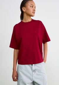 Tricou din bumbac burgund cu mânecă scurtă și guler rotund, având un mic accent cu logo pe piept, stilizat cu pantaloni lejeri din denim gri deschis.