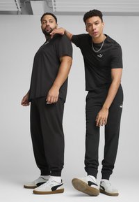 Twee mannen dragen zwarte PUMA T-shirts en bijpassende zwarte joggers. Beide hebben witte PUMA sneakers met zwarte accenten. Neutrale achtergrond.