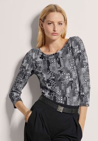 Langärmliges Oberteil in Schwarz-Weiß-Muster mit Rundhalsausschnitt, kombiniert mit einem schwarzen Gürtel mit einer metallischen "M"-Schnalle. Glatte Stoffoberfläche.