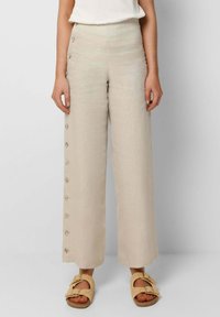 Kvinna som bär högmidjade vida beige byxor med knappar längs ena sidan, i kombination med ljusbeiga sandaler med öppen tå och spännen.