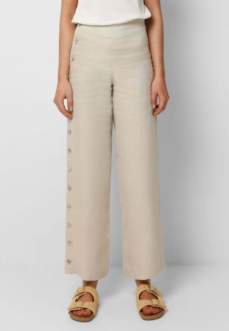 Kvinna som bär högmidjade vida beige byxor med knappar längs ena sidan, i kombination med ljusbeiga sandaler med öppen tå och spännen.