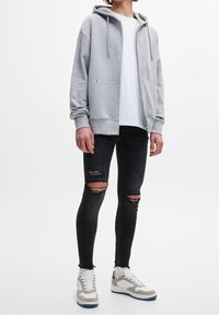 Persona con una sudadera gris con cremallera, camiseta blanca, jeans ajustados negros desgastados y zapatillas blancas con detalles grises, de pie contra un fondo blanco.