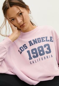 Ružový sweatshir s nápisom "LOS ANGELES 1983 CALIFORNIA" v námorníckej modrej farbe. Má okrúhly výstrih, rebrované manžety, mäkkú textúru tkaniny a voľný strih.