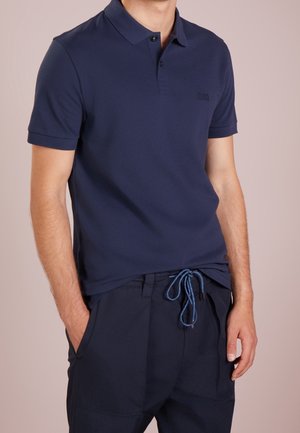 Torso masculin portant un polo à manches courtes bleu marine foncé et un pantalon bleu marine assorti avec une taille à cordon, mains partiellement dans les poches.