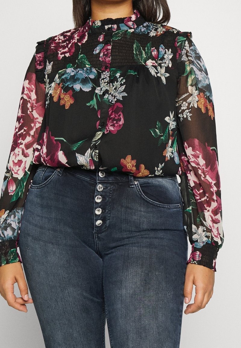 Femme portant un chemisier noir à manches longues à motif floral avec un col à volants et un jean bleu foncé taille haute avec cinq boutons argentés.