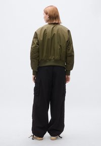 Χακί πράσινο μπουφάν τύπου μπόμπερ με ριμπ τελείωμα και μανσέτες, συνδυασμένο με χαλαρό μαύρο παντελόνι cargo με πλαϊνές τσέπες και μανσέτες με κορδόνι.