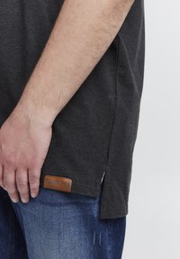 Tmavošedé tričko s krátkym rukávom so bočnými rozšíreniami a hnedou koženou etiketou. Nosené s svetlomodrými džínsami, ktoré zdôrazňujú bavlnenú textúru a ležérny strih.