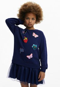 Felpa blu navy con toppe ricamate colorate, tra cui farfalle e una fragola, abbinata a una gonna in tulle a strati.