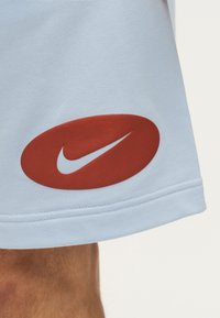 Ανοιχτό μπλε ύφασμα με ένα κόκκινο οβάλ που περιέχει το λευκό λογότυπο Nike swoosh κοντά στο τελείωμα πάνω από το γόνατο ενός ατόμου.