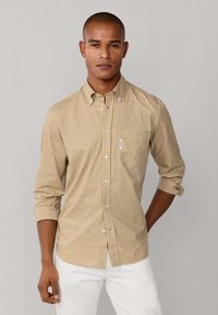 Camisa de botones a cuadros en tela de algodón marrón y blanca, con mangas largas, un bolsillo y puños enrollados. Llevada con pantalones blancos.