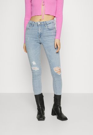 Mujer con jeans pitillo desgastados de mezclilla azul claro, un top rosa corto de manga larga y botines negros de tacón, de pie frente a un fondo liso.