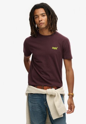 Camiseta de algodón color marrón con cuello redondo y mangas cortas, que presenta un pequeño logo amarillo en el lado izquierdo del pecho. Combinada con unos jeans azules y un suéter crema atado a la cintura.