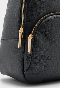 Melna teksturēta mugursoma ar zelta toņa rāvējslēdzējiem un vilkšanas cilpām, izliekta forma un priekšējā kabata papildu uzglabāšanai.