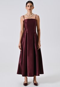 Robe en satin bourgogne avec un corsage ajusté, des fines bretelles et une jupe évasée longueur sol. Portée avec un collier de perles et des chaussures assorties.