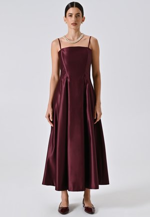 Abito di raso bordeaux con corpino aderente, spalline sottili e gonna a flusso lunga fino al pavimento. Indossato con una collana di perle e scarpe abbinate.