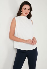 Fehér, ujjatlan felső, redőzött mintázattal, kerek dekoltázzsal és puha anyagból készült, sötét, testhezálló nadrággal párosítva. Egyszínű háttér előtt.