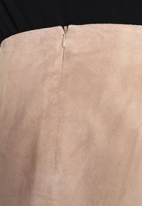 El tejido de gamuza en tono marrón claro tiene una textura suave y cuenta con una sección superior negra y un discreto detalle de cremallera en la parte de atrás, fusionando colores de manera armoniosa.