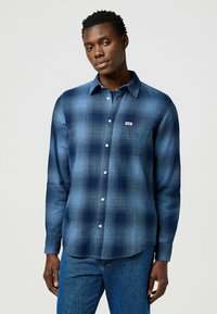 Camisa de cuadros azul de manga larga con botones blancos y un bolsillo en el pecho. Hecha de un material suave, presenta un corte relajado y un dobladillo redondeado.
