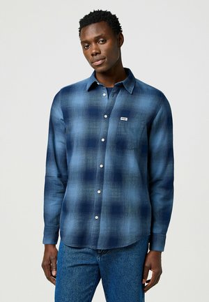 Camisa de cuadros azul de manga larga con botones blancos y un bolsillo en el pecho. Hecha de un material suave, presenta un corte relajado y un dobladillo redondeado.