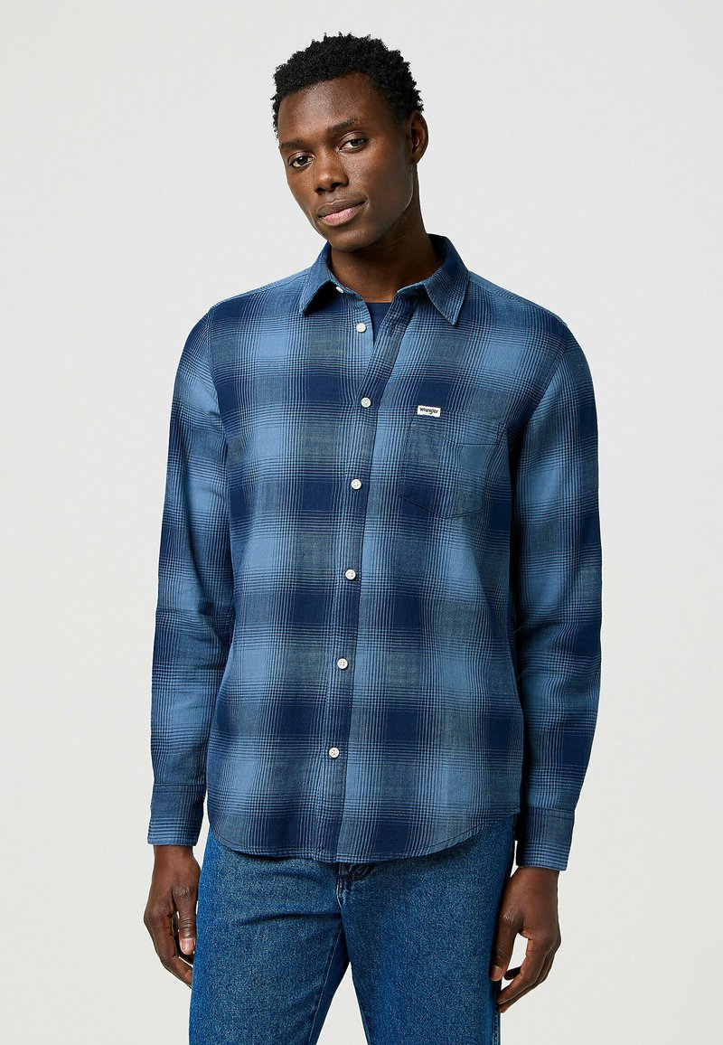 Camisa de cuadros azul de manga larga con botones blancos y un bolsillo en el pecho. Hecha de un material suave, presenta un corte relajado y un dobladillo redondeado.