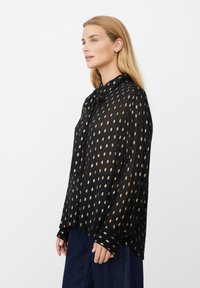 Schwarze transparente Bluse mit goldfarbenem Punktemuster, durchgehender Knopfverschluss, Kragen mit Bindedetail, langen Ärmeln und lockerer Passform. Texturierter Stoff.