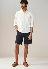 Chemise blanche à manches longues avec col boutonné, en tissu texturé ; associée à un short foncé et des sandales brunes avec des boucles pour les orteils.