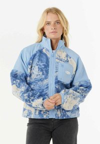 Blonde vrouw in een blauwe en witte geblokte fleecejack met hoge kraag en ritssluiting, staande tegen een effen achtergrond.