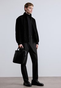 Čierna semišová bombardérska bunda, čierny rolák, čierne šité nohavice a čierne kožené topánky. Drží čiernu koženú aktovku.