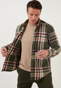 Camisa de franela en verde con un patrón de cuadros en rojo, blanco y marrón. Presenta un cierre frontal con botones y mangas largas, combinada con una camiseta interior beige.
