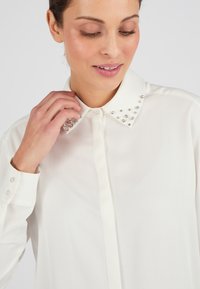 Blusa blanca con un cuello clásico adornado con tachuelas plateadas. Tejido liso y liviano con puños de botón y corte recto.