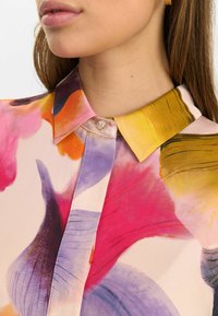 Färgglad silkesblus med krage, stora blommönster i rosa, lila och orange, med en knapp vid kragen och en mjuk textur.