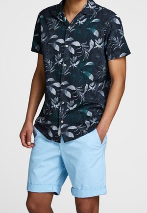Man draagt een donkerblauw overhemd met korte mouwen met witte en groene bladvormen en lichtblauwe opgerolde shorts.